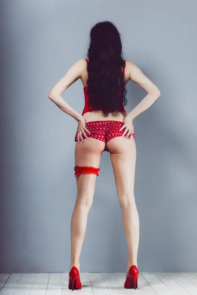 Sexy chica morena mira hacia atrás mientras está de pie y sosteniendo su culo en bragas rojas y tacones de corsé con liga en el estudio — Foto de Stock