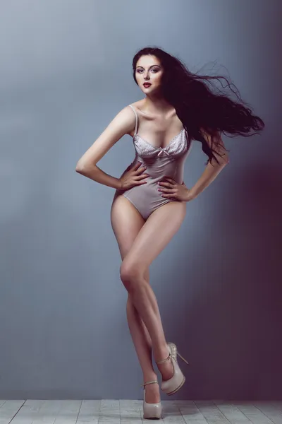 Sexy brunette girl in a transparent body beige heels with a beautiful body and long legs and hair developing on a wind in studio on white wooden floor and gray walls