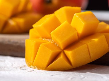 Ahşap kesim tahtasının üzerinde beyaz bir tabakta kesilmiş taze mango meyvesi ve gri masa arkasında yapraklar..