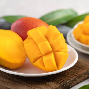 Ahşap kesim tahtasının üzerinde beyaz bir tabakta kesilmiş taze mango meyvesi ve gri masa arkasında yapraklar..
