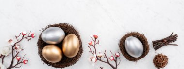 Yuvasında altın ve gümüş Paskalya yumurtaları, parlak mermer masa arkasında beyaz erik çiçekleri olan tasarım konsepti..