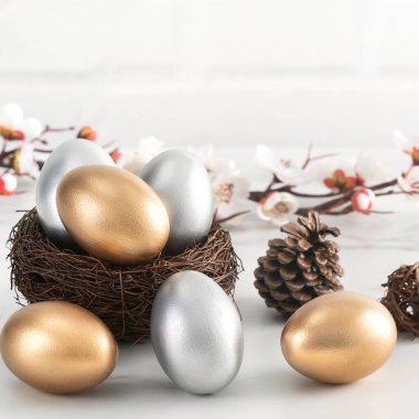 Yuvadaki altın ve gümüş Paskalya yumurtalarını parlak beyaz ahşap masa zemininde beyaz erik çiçeğiyle kapat.