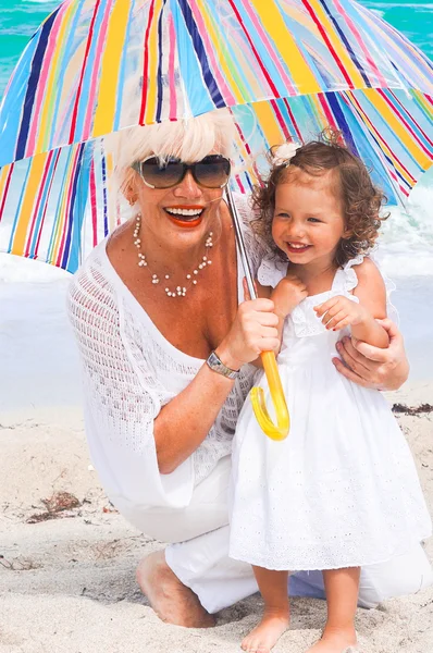 Happy grandmother and granddaughter under umbrella at the beach — Stock Photo, Image