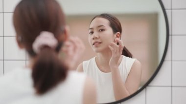 Closeup portrait of smiling young beautiful asian woman looking at mirror touching her skin enjoying treatment for dry skin. 
