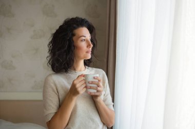 Çekici genç bir kadın elinde bir fincan çayla pencereye bakıyor. Sıcak ev atmosferi.