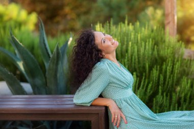 Kıvırcık saçlı, yaz elbisesi giymiş, gülümseyen esmer kadın. Dışarıda poz veren mutlu kadın..