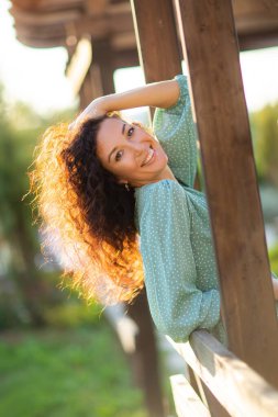 Kıvırcık saçlı, yaz elbisesi giymiş, gülümseyen esmer kadın. Dışarıda poz veren mutlu kadın..