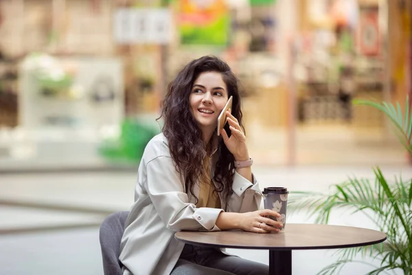 Genç ponpon kız akıllı telefon kullanıyor ve kahve içiyor kağıt bardakta. Cafe 'de oturan şık bir kadın..
