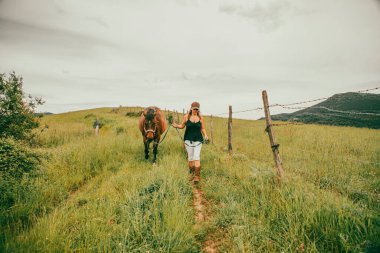 Amazon kadını elinde kahverengi bir atla kırsalda yürüyor. Kovboy kadın kahverengi şapka, siyah tişört, beyaz pantolon ve binici botları giyiyor. Kırsal alanda arka planda evleri olan yeşil manzara görülüyor.