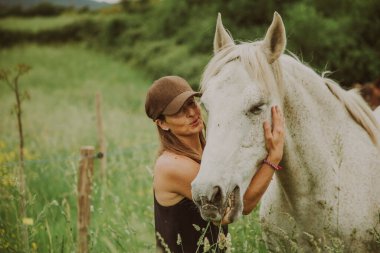 Çayırda uzun otlu bir kadın beyaz bir atı okşuyor..