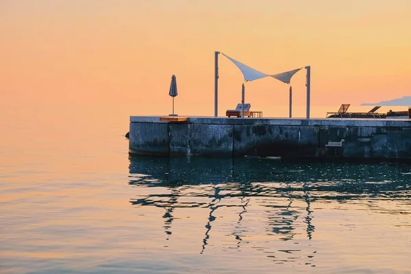 Fin de la temporada de vacaciones en la playa, vista panorámica a las siluetas del pabellón de lujo vacío montado con sombrilla cerrada y tumbona al atardecer en reflejos de agua naranja rosada en la costa mediterránea. — Foto de Stock