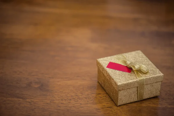 Золотая подарочная коробка с бантом и небольшой карточкой на деревянном столе — стоковое фото