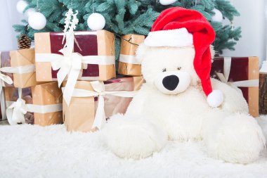 Noel Baba şapkası takan oyuncak ayı hediye kutularıyla bir Noel ağacının altında oturuyor..