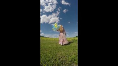 Güzel genç bir kadın, güneşli bir günde elinde balonlarla bir çiçek tarlasında yürüyor. Dikiz aynası. Balonlarla parkta yürüyorum. 4K. 