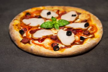Appetizing pizza with tomatoes, bacon, black olives, basil and cheese on black concrete background. Top view.