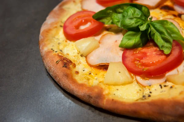 Appetizing pizza with pineapple, tomatoes, chicken meat, basil and cheese on black concrete background. Hawaiian pizza. Top view.