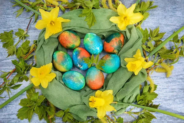 Primo piano di uova di Pasqua in un nido decorativo fatto di rami verdi, narcisi gialli e un tovagliolo verde su sfondo blu. Vista dall'alto. — Foto Stock