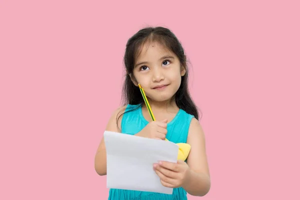 Portrait with copy space empty place of thoughtful little Asian girl having paper and pencil in hands looking up, writer waiting for muse, isolated on pink background