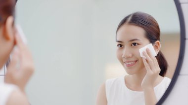 Mutlu Asyalı kız aynaya bakıyor, elinde pamuk tutuyordu, yüzüne yüz silme uyguluyor, makyaj malzemelerini temizliyordu..
