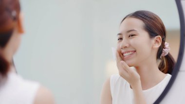 Happy Asian girl looking in to mirror, holding cotton pad, applying facial wipe on her face, removing makeup.
