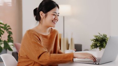 Genç modern Asyalı kadın evde dizüstü bilgisayarla oturma odasında çalışıyor..