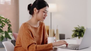Genç modern Asyalı kadın evde dizüstü bilgisayarla oturma odasında çalışıyor..