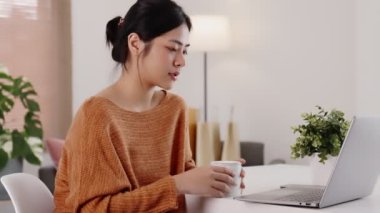 Genç modern Asyalı kadın evde dizüstü bilgisayarla oturma odasında çalışıyor..