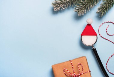 Kırmızı Noel Baba şapkalı tıbbi steteskop hediye kutusu ve açık mavi arka planda şenlik dekorasyonu. Yaratıcı tıbbi Noel ve yeni yıl tebrik kartı. Üst görünüm, düz görünüm, kopyalama alanı