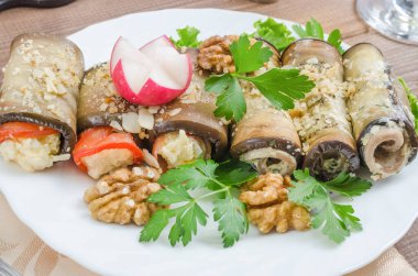 Peynirle doldurulmuş sağlıklı patlıcan ruloları, siyah ahşap arka planda beyaz bir tabakta yakın plan sebze ve otlarla servis edilir.