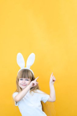 Sarı arka planda tavşan kulaklı neşeli bir kız. Komik mutlu çocuk boş bir yeri işaret ediyor. metin ve model için boşluk kopyala
