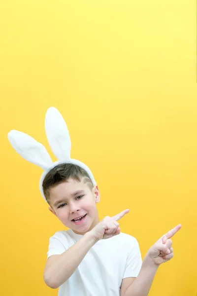 一个快乐的男孩 头上戴着兔子耳朵 背景是黄色的 有趣而快乐的孩子把手指指向一个空旷的空间 复制空间为文本 复活节的概念 — 图库照片