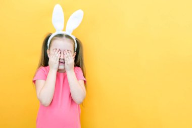 Sarı arka planda tavşan kulaklı neşeli bir kız. Komik, çılgın, mutlu çocuk. Paskalya çocuğu. Elleriyle gözlerini kapatıyor. Promosyon ürünleri. metin ve düzen için boşluk kopyala