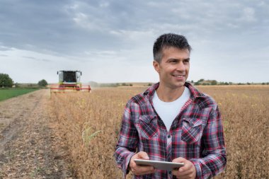 Erkek tarım uzmanı gülümsüyor ve tarımda tablet modern teknolojiyi kullanıyor. Soya tarlasında dikilen ve arka planda çalışan bir adam..
