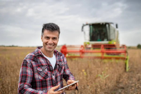 男性農学者は農業のタブレット現代技術を笑顔と使用しています 背景に作業をしている収穫者と大豆畑に立つ男 — ストック写真