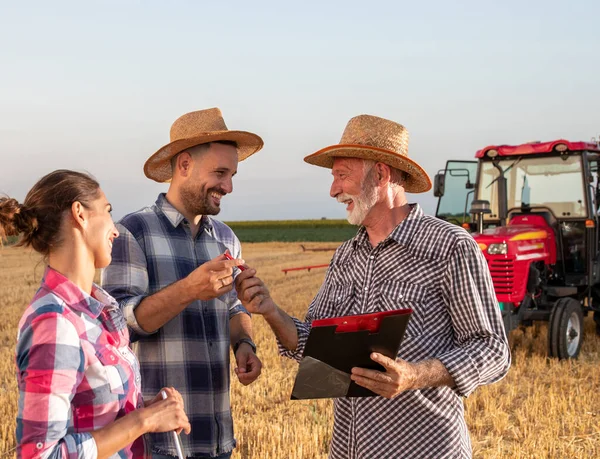 若い農家のペンを与えるクリップボードを保持する高齢農業学者 トラクターを背景に小麦畑に立ちながら保険販売担当者と商談を行う2人の農家 — ストック写真