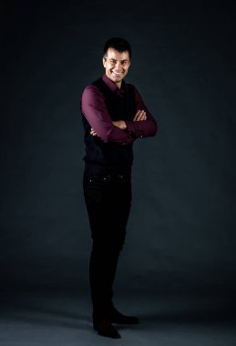 Stüdyoda siyah arka planda kolları çapraz duran yakışıklı beyaz adam zarif siyah pantolon ve yelek giyiyor.