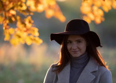 Sonbaharda günbatımında ormanda poz veren şapkalı ve ceketli güzel kızıl saçlı kızın portresi.