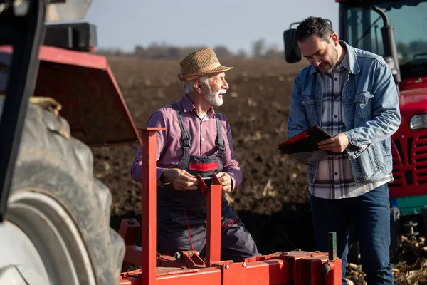 Senior Landwirt Arbeitet Ausrüstung Des Traktors Während Jüngerer Mann Notizen — Stockfoto