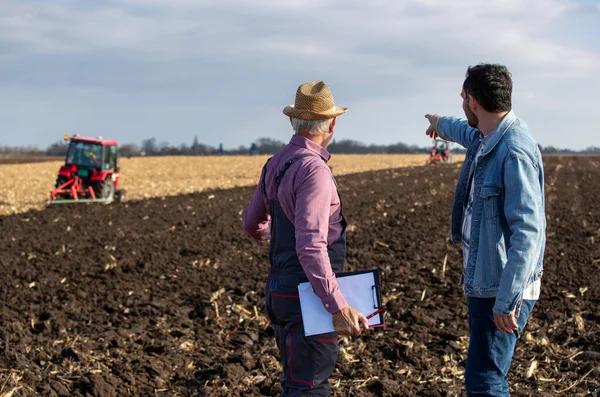 Dwóch Farmerów Stojących Polu Rozglądających Się Daleko Ciągnik Pracujący Tle — Zdjęcie stockowe