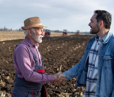 Two farmers senior worker and younger engineer shaking hands in front of tractors in field in autumn time
