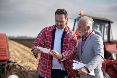 Mid adult farmer and his insurance agent reading documents on agricultural field.