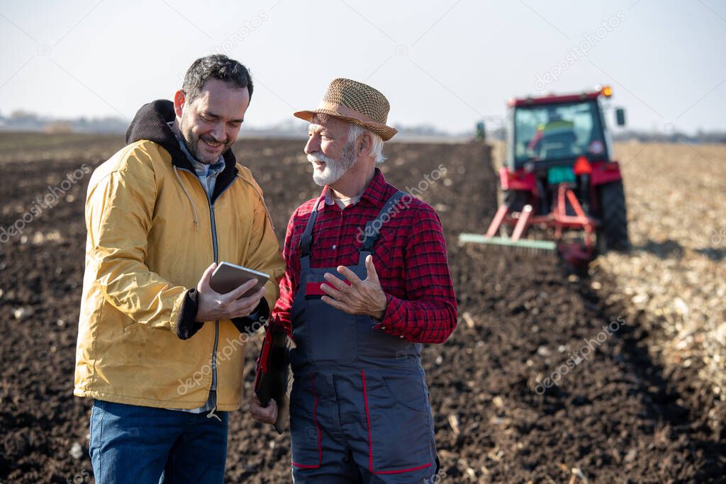 Two farmers standing in field in autumn, holding tablet and talking while tractor working in background