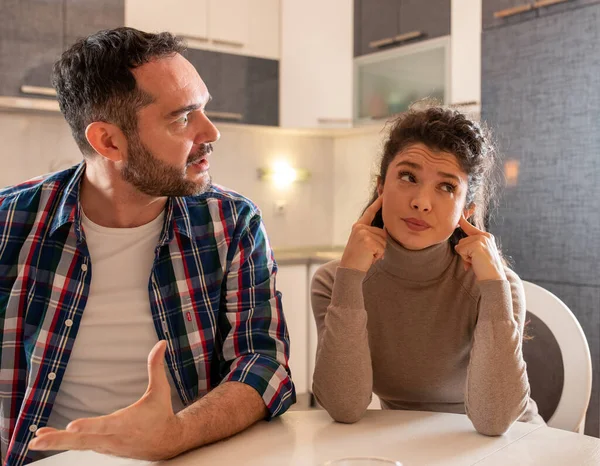 Husband and wife having argument at dining table at home. Man explaining something with dominant posture and woman holding fingers in ears and having bored expression