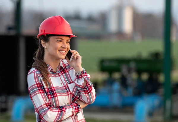 農業分野の灌漑システムプラントの前に立つヘルメットと彼女の電話で話す若い女性エンジニアを笑顔 — ストック写真