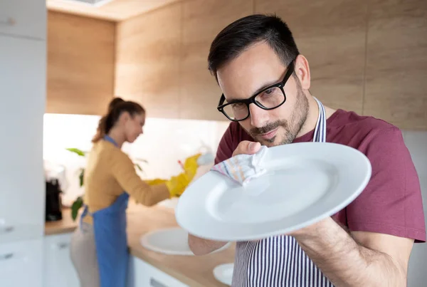 厨房里拿围裙的男人用布擦拭盘子 而后面的女人则洗碗 — 图库照片