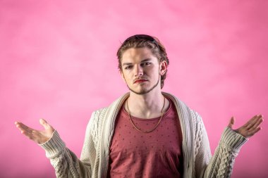 Yüzünde sakalı ve makyajı olan genç bir adamın portresi. Kollarını kaldırıp pembe arka plana doğru erkekçe bakıyor. Stüdyo çekimi.