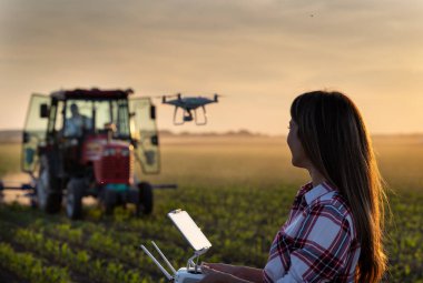 Güzel bir çiftçi kadın, sonbaharda mısır tarlasında traktörün önünde uzaktan kumandalı drone kullanıyor.