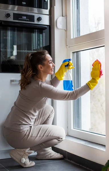 戴橡胶防护手套的年轻女子用细纤维布擦窗户 另一只手拿着喷雾器 — 图库照片