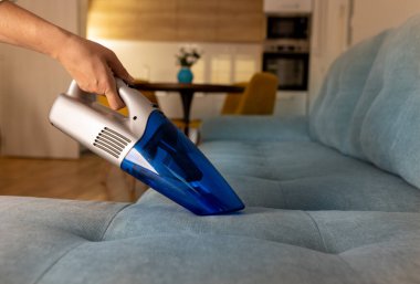 Oturma odasında, arka planda mutfak ve yemek masası olan oturma odasındaki kanepeden alınan saç kırıntılarını temizleyen kablosuz elektrikli süpürge tutan eldivenlerle kadın elini kapat. Oda servisi, temizlik, hijyen ve bakım konsepti