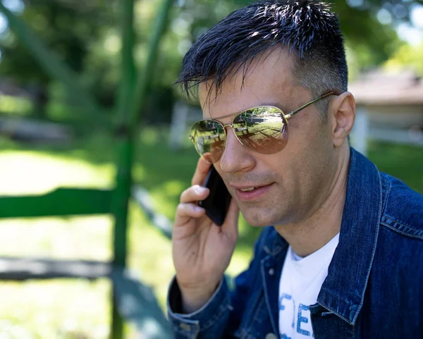 Handsome man in jeans jacket wearing sunglasses in park and talking on mobile phone